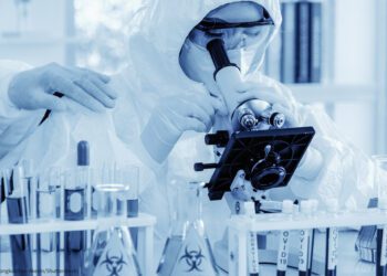 scientist in biohazard protection clothing analyzing covid 19 sample with microscope and holding coronavirus covid 19 blood sample tube on hand in laboratory, coronavirus covid 19 vaccine research
By (© Mongkolchon Akesin/Shutterstock)